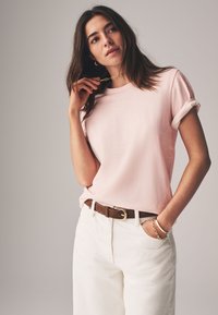 Rosa t-shirt med korta ärmar och uppvikta ärmslut, kombinerad med vita byxor med hög midja. Outfiten har ett brunt bälte och ett guldigt kedjebracelet.