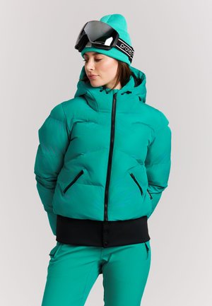 Vrouw in turquoise skijas en broek die een bijpassende hoed en zwarte skibril op haar voorhoofd draagt, kijkt opzij.