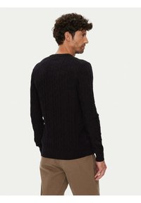Schwarzer Pullover mit Zopfmuster, langen Ärmeln, gerippten Bündchen und Saum, mit strukturiertem Muster. Getragen mit beigen Hosen.