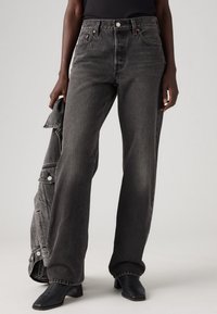 Zwarte denim wijd-zijde jeans met een hoge taille, voorzien van koperen hardware en een subtiele vervaagde textuur. Model houdt een bijpassende jas vast.