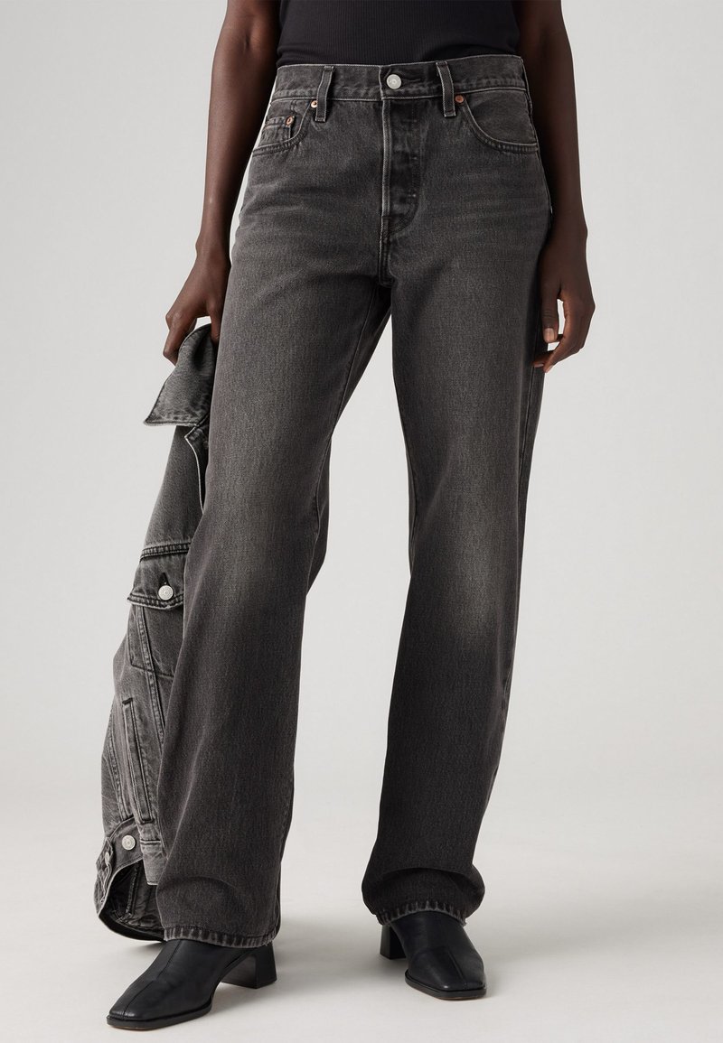 Sorte denim bred-leg jeans med høy midje, med kobberfargede detaljer og en subtil falming. Modellen holder en matchende jakke.