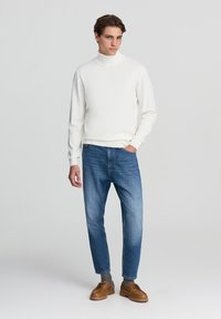 Maglione bianco con colletto a dolcevita e polsini a coste, abbinato a jeans azzurri e scarpe marroni con lacci, il tutto su uno sfondo neutro.