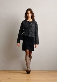 Veste cropped en tweed noir avec des boutons argentés, associée à une robe noire et des bottes beige jusqu'au genou, posée sur un sol en bois clair.