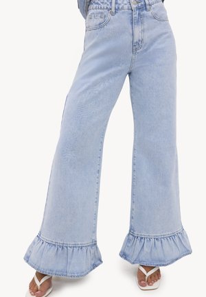 Jean bleu clair à jambes larges avec des ourlets volantés porté avec des sandales blanches type tongs sur une personne debout devant un fond uni.