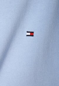 Jasnoniebieski sweter bawełniany z małym haftowanym logo w czerwonych, białych i czarnych akcentach na lewej stronie klatki piersiowej.