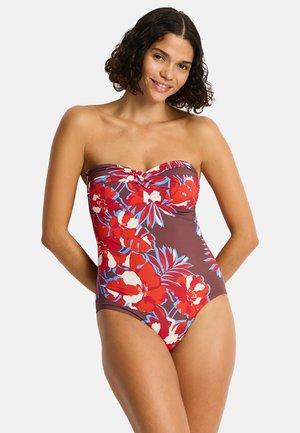 Femme portant un maillot de bain une pièce sans bretelles avec un imprimé floral rouge, blanc et bleu sur fond marron, souriante, les mains derrière le dos.
