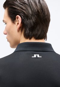 Νεαρό άτομο με κοντά σκουρόχρωμα μαλλιά, που φοράει μαύρο πουκάμισο με γιακά, στο πίσω μέρος κοντά στον γιακά εμφανίζεται μικρό λευκό στυλιζαρισμένο λογότυπο.