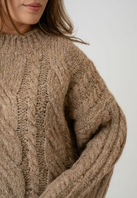 Bežani pleteni džemper s debelom čipkastom šablonom, okruglim izrezom i spuštenim ramenima. Tekstura izgleda gusta i udobna.