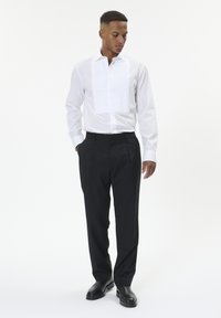 Valge pluus, millel on voltidega esiosa, pikad varrukad, koos mustade ülikondipükstega, millel on täpselt istuv lõige ja sile kangas.