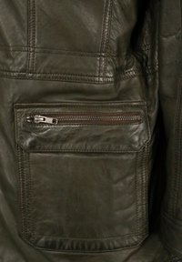 Dunkelgrüner Lederjackenbereich mit einer Reißverschlusstasche, strukturiertem Finish, sichtbaren Nähten und einem horizontalen Nahtdetail.