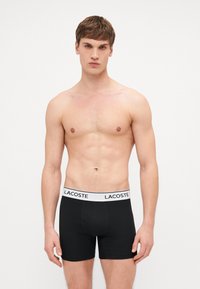 Fekete boxeralsó fehér Lacoste logós derékkal, sima anyagból készült, testhezálló fazonnal és combközép hosszúsággal.