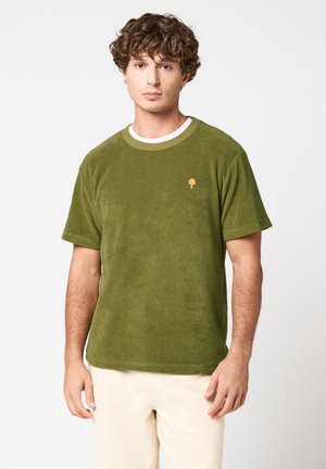 Jeune homme aux cheveux bouclés portant une chemise verte à manches courtes avec un petit logo d'arbre orange et un pantalon de couleur claire sur un fond blanc.