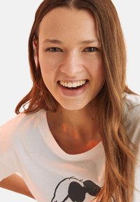 T-shirt bianco in cotone con maniche corte, caratterizzato da un design grafico nero. Il modello ha lunghi capelli castani mossi e un sorriso luminoso.