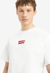 Weißes Baumwoll-T-Shirt mit einem roten bestickten Levi's-Logo in einem klassischen Patch-Design. Kurzärmelig und mit lockerer Passform.