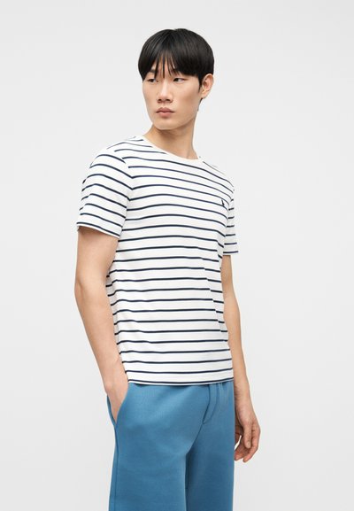 Νέος άνδρας με μαύρα μαλλιά, φορώντας ένα λευκό και ναυτικό μπλε ριγέ κοντομάνικο πουκάμισο και μπλε παντελόνι, στέκεται μπροστά σε απλό φωτεινό φόντο.