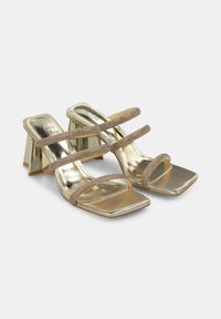 Goldene metallische Sandalen mit Absatz, zwei überkreuzten Riemen, quadratischer Zehenpartie und einem einzigartigen geometrischen Blockabsatz. Glatte Textur mit schimmernden Akzenten.