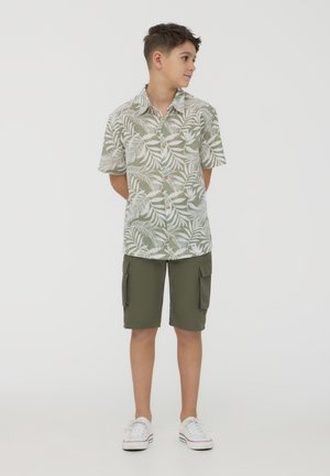 Maglietta a maniche corte con motivo di foglie verdi e bianche, bottoni sul davanti; abbinata a pantaloni cargo verde oliva e sneakers bianche.