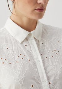 Λευκό πουκάμισο με κέντημα και γιακά. Διαθέτει κυκλικά κοψίματα και περίπλοκα σχέδια, κατασκευασμένο από ελαφρύ ύφασμα με κουμπιά.