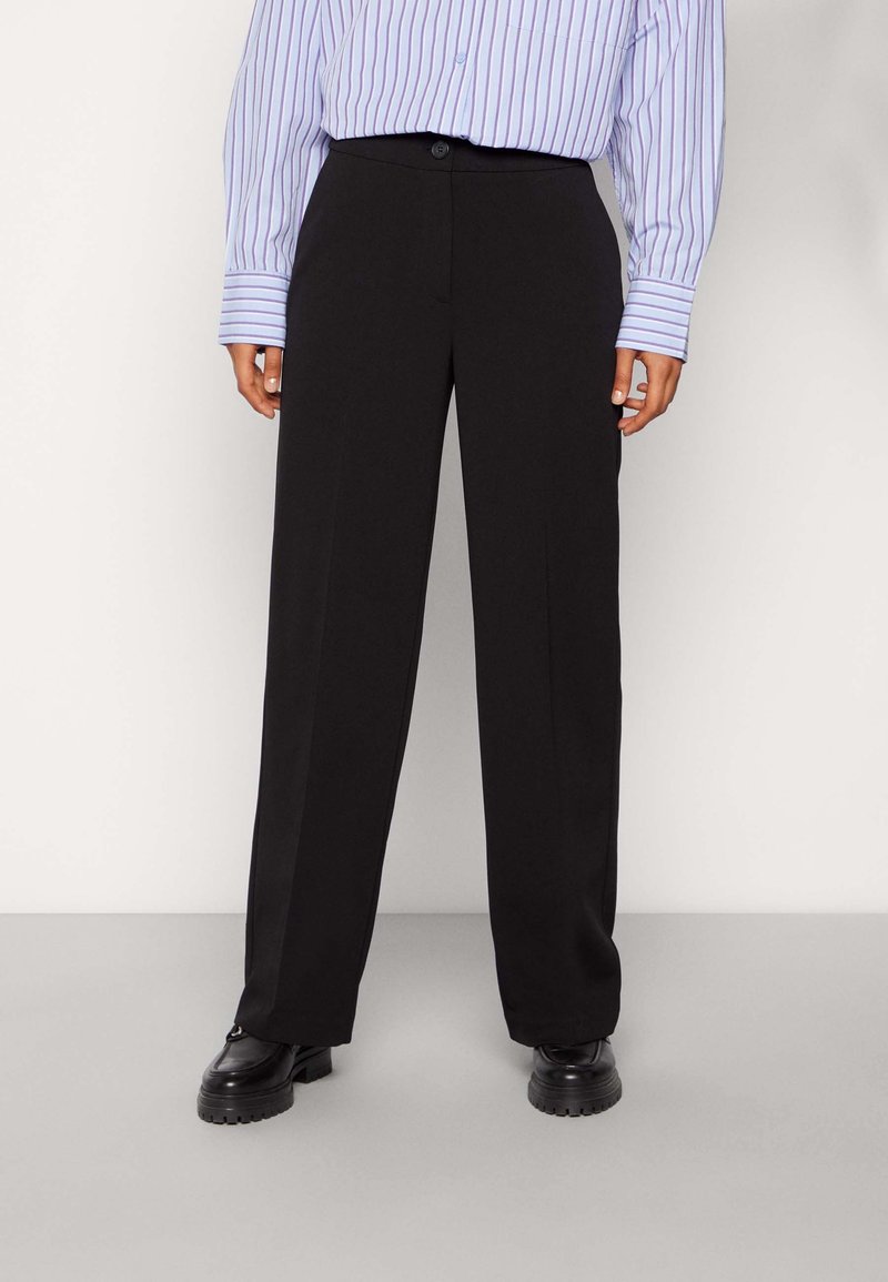 Personne portant un pantalon noir à jambes larges, des chaussures noires à semelle épaisse et une chemise à rayures bleues et blanches à manches longues.