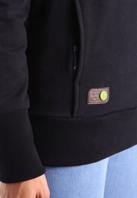 En närbild av en person som bär en svart sweatshirt med ribbad mudd och en brun lapp med en grön knapp nära fickan, i kombination med ljusblå jeans.