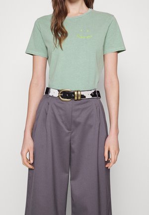 Groen katoenen T-shirt met gebogen gele tekst, gecombineerd met grijze wijde broekspijpen en een zwart-witte riem met een metalen gesp.