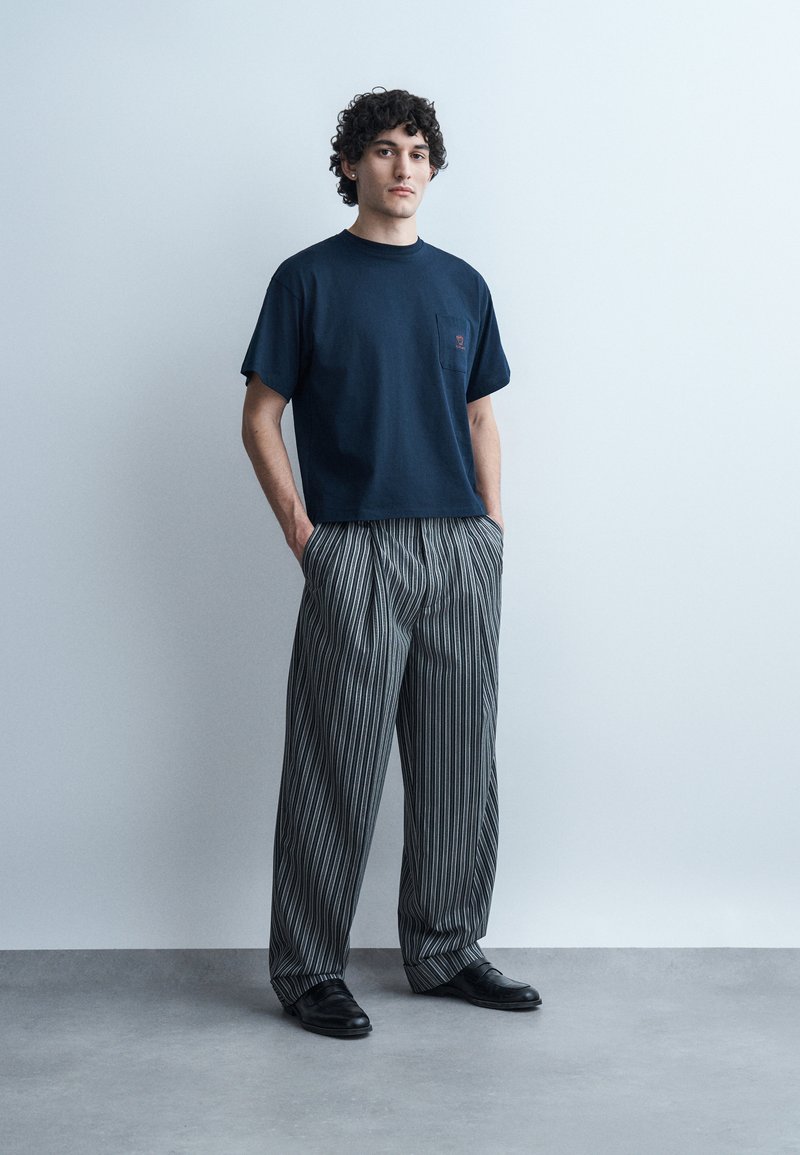 Giovane uomo con capelli ricci in piedi con le mani nelle tasche, indossa una maglietta blu navy e pantaloni larghi a righe bianche e nere su sfondo semplice.