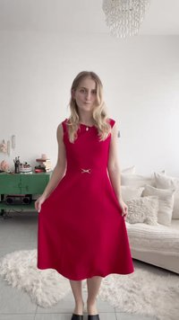 Femme aux cheveux blonds portant une robe midi rouge sans manches, debout dans un salon lumineux avec une armoire verte et un canapé beige en arrière-plan.