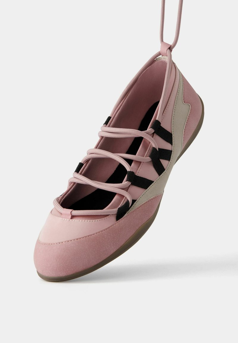 Zapatilla deportiva de color rosa claro con una parte superior suave y texturizada, que presenta cordones elásticos negros y detalles en gris. Punta redondeada y suela flexible.