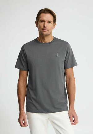 Uomo con t-shirt grigia a maniche corte con piccolo logo bianco di un golfista, che indossa pantaloni bianchi, in piedi contro uno sfondo chiaro uniforme.