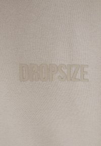 Sweatshirt beige avec un logo "DROPSIZE" en relief et texturé au centre. Le matériau semble doux et tricoté avec des variations de tons subtiles.