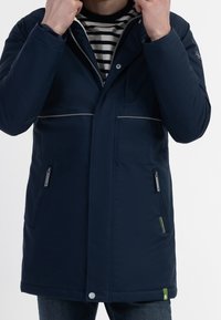 Navy blue winter coat with a zip and snap closure, featuring two side pockets, contrasting grey piping, and a green accent at the hem.