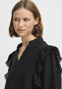Blusa nera leggera con tessuto texturizzato. Presenta un scollo a V e maniche a sbuffo con bordo in pizzo. Design elegante e semplice.