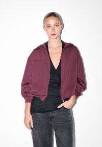 Mladá žena s previazanými vlasmi, oblečená v marónovej mikine na zips cez čierny top s bodkami a tmavé džínsy, pózuje s jednou rukou vo vrecku.