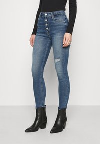 Mujer vistiendo jeans ajustados de cintura alta en azul con cierre de botones y rasgadura en la rodilla, combinados con botines negros de tacón y una blusa negra de manga larga.