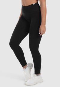 Leggings neri a vita alta realizzati in tessuto elastico, con una texture liscia e design senza cuciture. Indossati con scarpe sportive bianche.