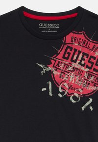 Svart T-skjorte i organisk bomull med rødt grafisk design med et skjold, teksten "GUESS" og "1981," samt rund hals med røde detaljer.