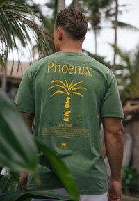 Zelené bavlněné tričko s žlutou grafikou palmy na zádech, s textem "Phoenix" nahoře a popisným textem níže.