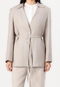 Beige Leinenmischung Blazer mit langen Ärmeln, Reverskragen, Gürtel in der Taille und zwei Knöpfen; glatte Textur und minimalistischer Stil.