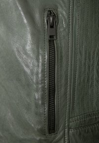 Olivengrüne Lederjacke mit strukturierter Oberfläche, seitlichem Reißverschluss mit schwarzem Metall und horizontalen Steppnähten entlang der Nähte.