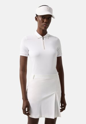 Frau trägt ein weißes Poloshirt mit kurzen Ärmeln, einen weißen Faltenrock und ein weißes Visier, steht vor einem schlichten hellgrauen Hintergrund.