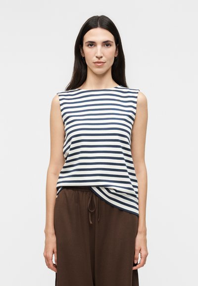 Femme aux longs cheveux foncés portant un haut sans manches rayé bleu marine et blanc et un pantalon marron ample, debout devant un fond clair uni.