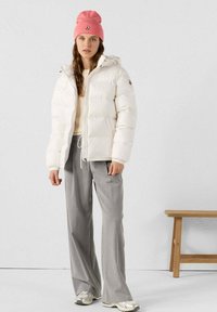 Witte donsjas met capuchon, met een gestructureerde afwerking, gedragen over een crème trui en grijze wijde broek. Een roze beanie voegt een kleuraccent toe.