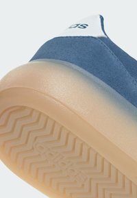 Plave zamšaste tenisice s prozirnom gumenom potplatom koja ima teksturirani uzorak u obliku cik-caka i bijeli naglasak na peti s natpisom "adidas."