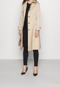 Beige trenchcoat med krage, knappstängning och axelklaffar. Kombinerad med svarta leggings och klackar, hållande en brun väska.