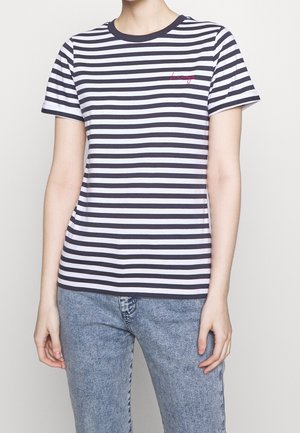 Osoba ubrana w czarno-białą, poziomo paskowaną koszulkę z krótkim rękawem, z małym czerwonym napisem na piersi oraz w niebieskie dżinsy.