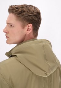Olivenfarvet hættejakke med en glat, blank overflade, der har en frontlynlås og subtile syninger, båret af en model set fra siden.