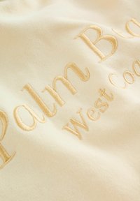 Krēmkrāsas audums ar zelta izšuvumu, uz kura rakstīts "Palm Beach West Coast", raksturots ar gludu tekstūru un ikdienišķu dizainu.