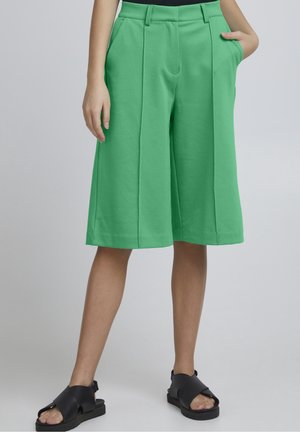 Personne portant un short vert ample arrivant aux genoux, des sandales noires à bouts ouverts, une main dans la poche et l’autre le long du corps.