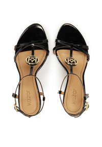 Černé lakované kožené sandály na podpatku s křížovými pásky, zlatým logem a béžovou stélkou. Hladká textura a otevřený design špičky.