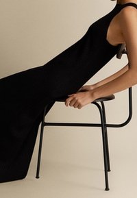 Robe noire en tricot côtelé au design sans manches, descendant jusqu'au sol, drapée sur une chaise en métal noir au design minimaliste.